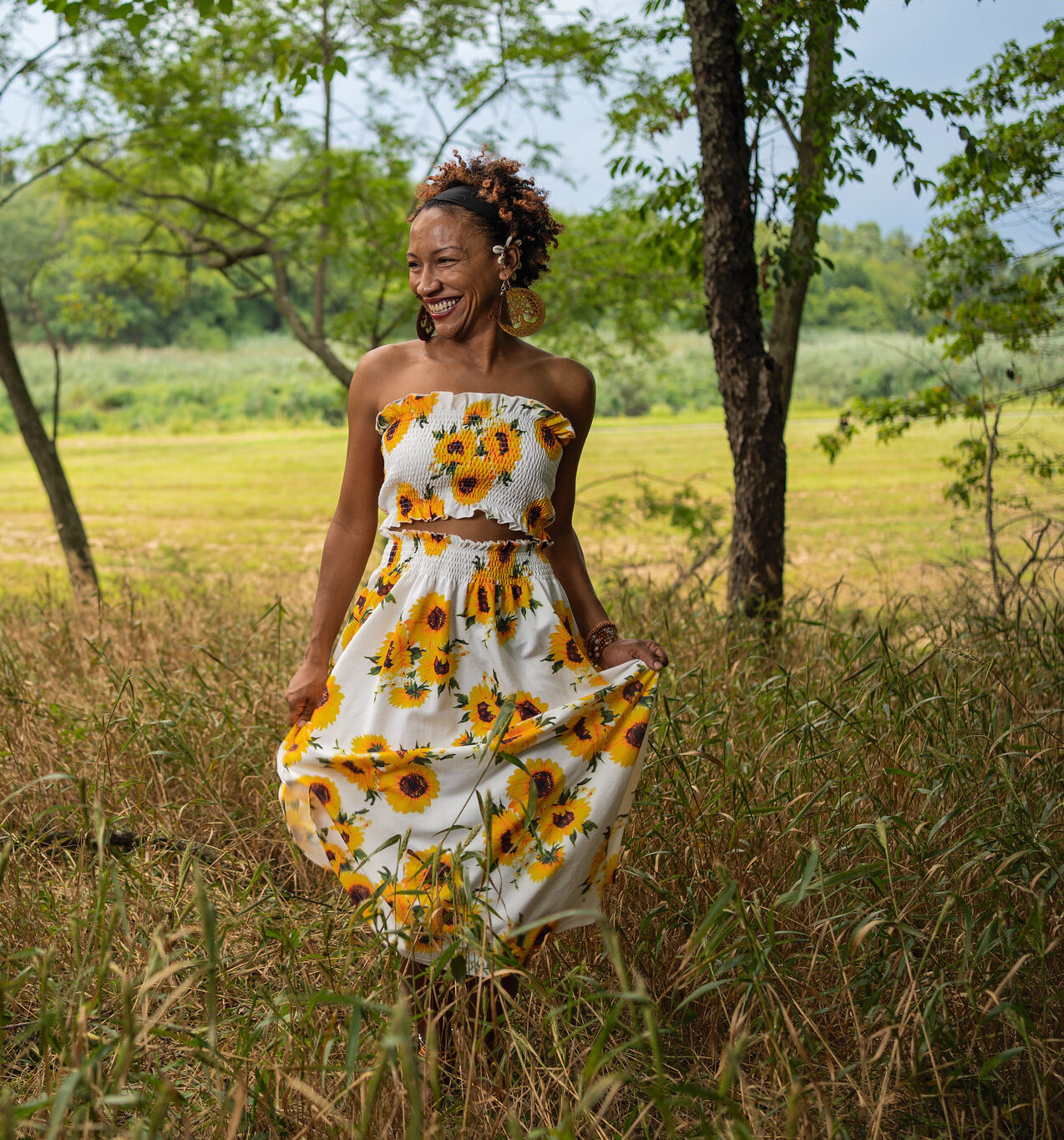 A Black woman in a sunflowers dress stands in a field of grassy vegetation, shaded by trees, with sunlight on the fields and forests behind her.
